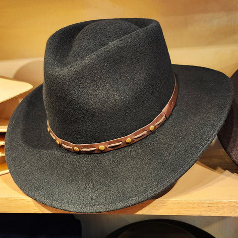 A black wool felt cowboy hat with a brown leather band decorated with brass studs, displayed on a wooden shelf.