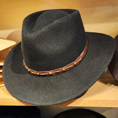 A black wool felt cowboy hat with a brown leather band decorated with brass studs, displayed on a wooden shelf.